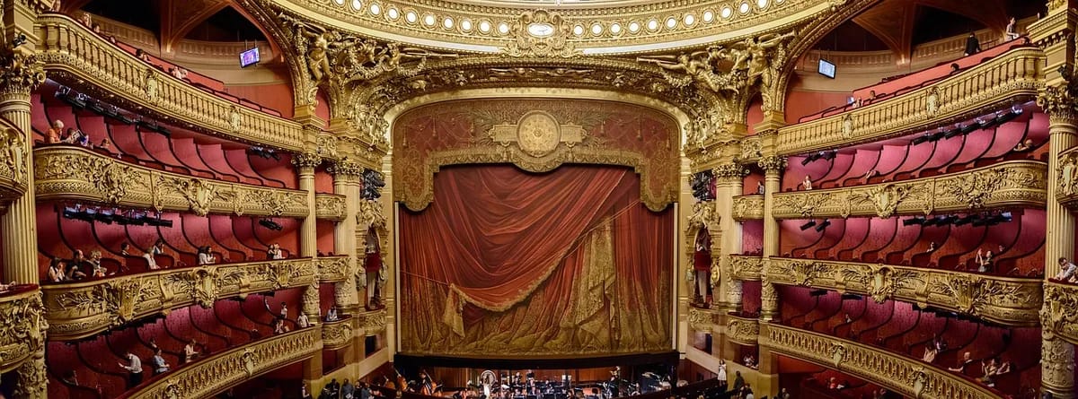 Der geschlossene rote Vorhang einer Theaterbühne, umrahmt von vergoldetem Stuck und den Rängen des Zuschauerraums